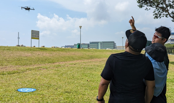 Three people looking up at a drone flying over a grassy field with buildings in the background.
